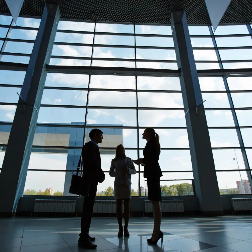 Trois professionnels d'affaires se tiennent devant de grandes fenêtres, discutant dans un environnement lumineux