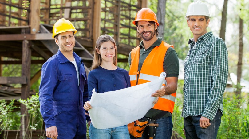 Trois personnes en casques de sécurité tenant un plan de construction, discutant d'un projet sur un site de travail