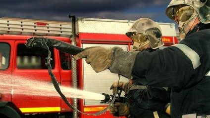 Pompiers en action, éteignant un incendie avec des tuyaux et des équipements de protection, sous un ciel fumant