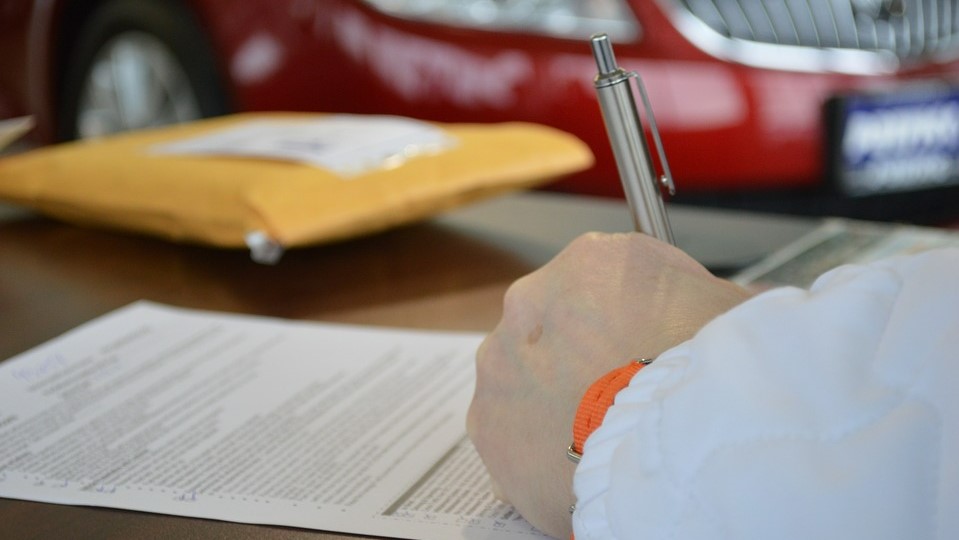 Une personne écrit sur du papier avec un stylo, tandis qu'une voiture rouge est garée à proximité