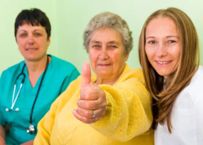 Une femme fait un signe de pouce levé à deux infirmières souriantes, exprimant sa gratitude et son soutien