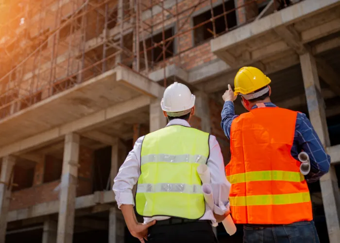 Deux ouvriers du bâtiment se tiennent devant un immeuble en construction, portant des casques de sécurité et des gilets réfléchissants