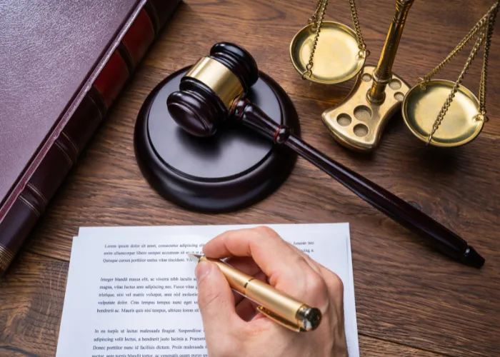 Main d'un avocat écrivant sur du papier, avec un maillet et des balances de la justice en arrière-plan